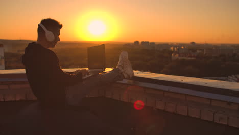 A-man-in-a-hoodie-sits-on-the-roof-and-listens-to-music-with-headphones-typing-on-a-laptop-keyboard.-Run-at-sunset.-Freelancer-works-at-sunset-with-a-laptop